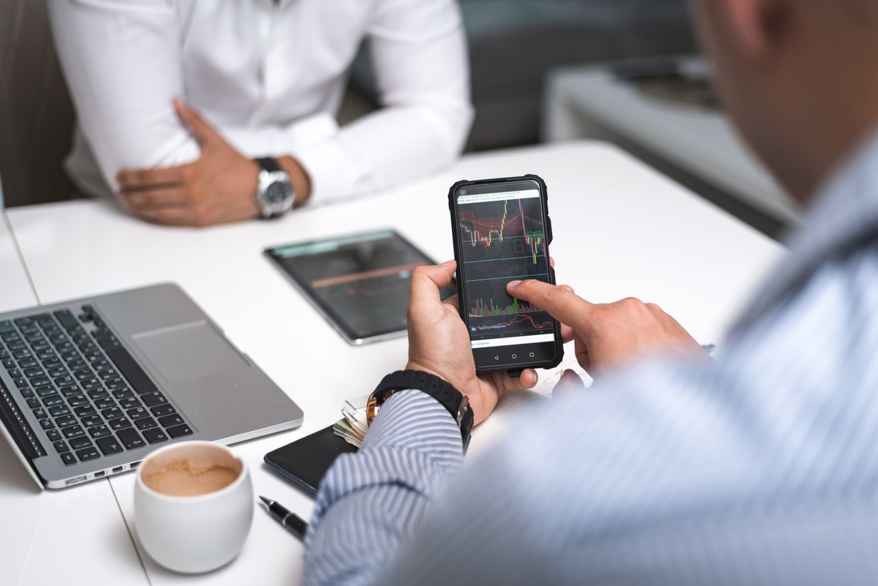 Home Two businessmen analyze stock market data using a smartphone and laptop in a modern office setting.